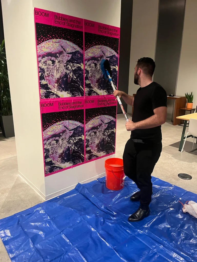 A man applies paste to four vibrant posters titled "BOOM: Bubbles and the End of Stagnation" on a wall. He stands on a blue tarp with a red bucket nearby. wheatpasting