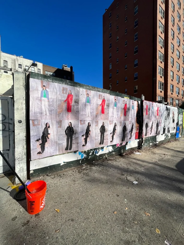 A series of fashion posters featuring various models wearing colorful outfits is displayed on a street wall. A red bucket and mop are nearby. Urban vibe. Wildposting