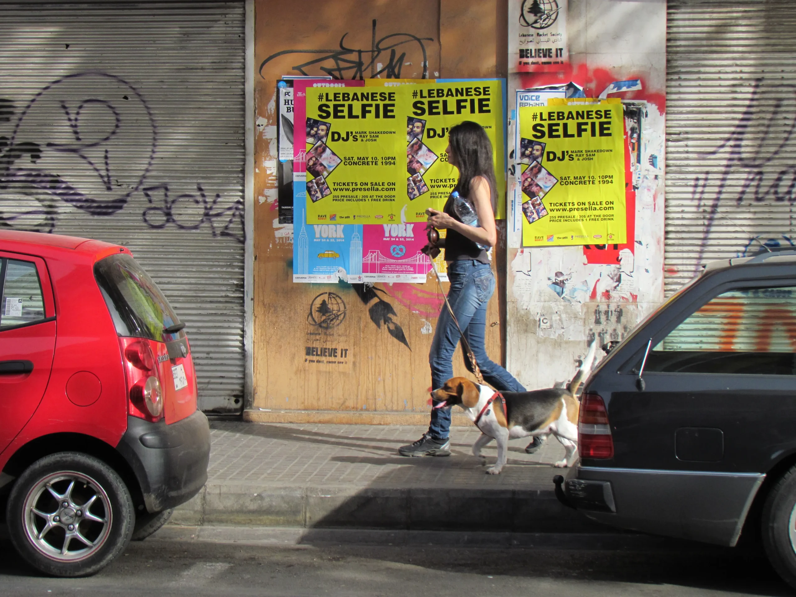 A woman walks her dog on a sunny street past vibrant posters for a Lebanese event on a graffiti-covered wall, flanked by red and black cars.