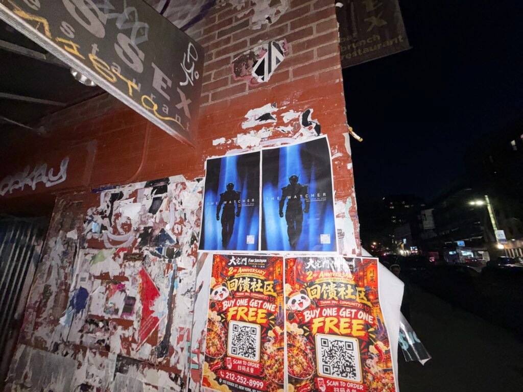 Two blue The Watcher posters installed side by side on a brick wall at night.