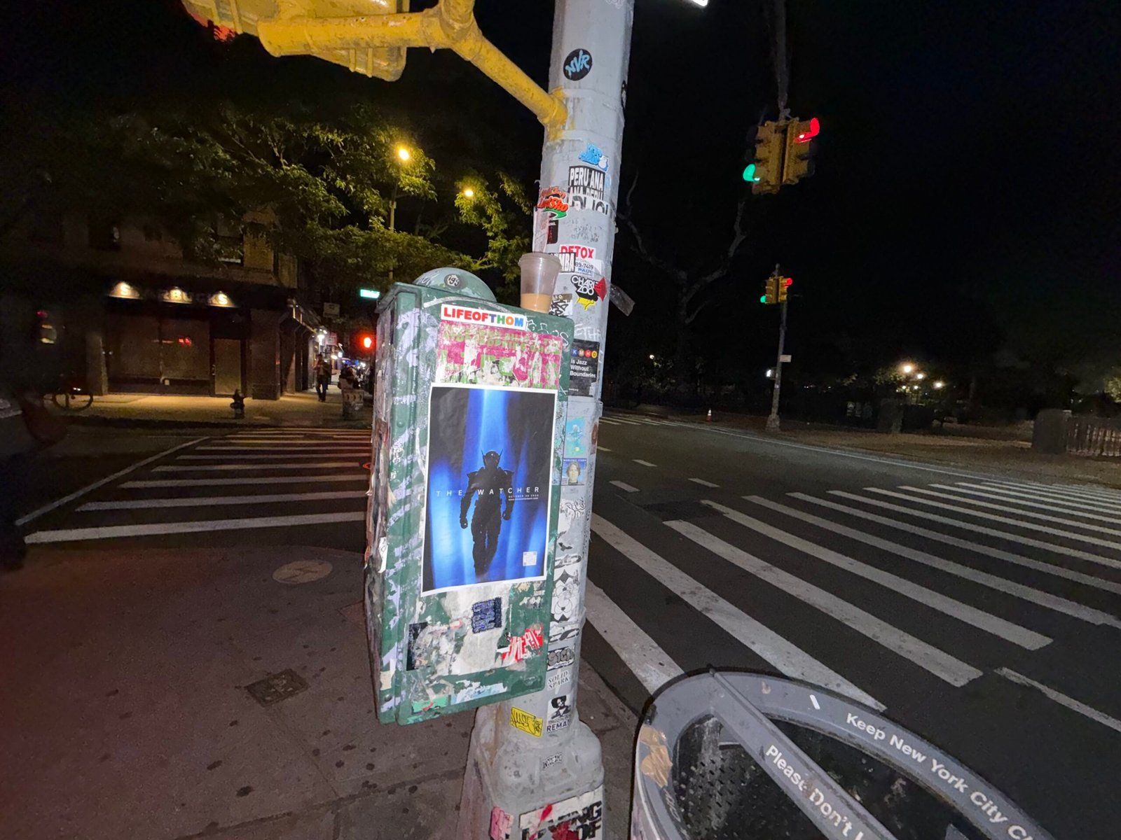 Single The Watcher poster beside a street-corner crosswalk at night.