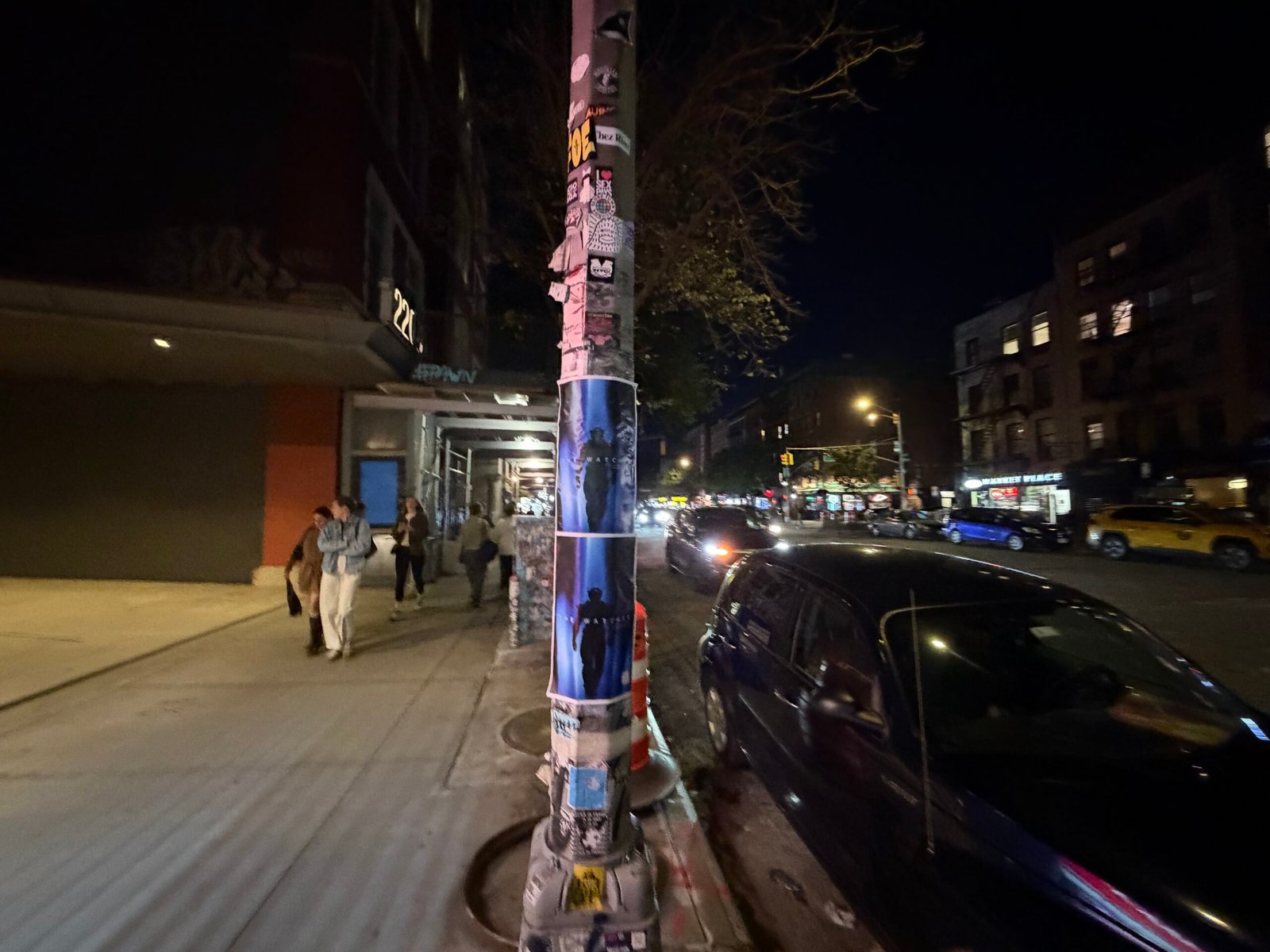Stacked The Watcher posters on a sidewalk pole with pedestrians and traffic nearby.
